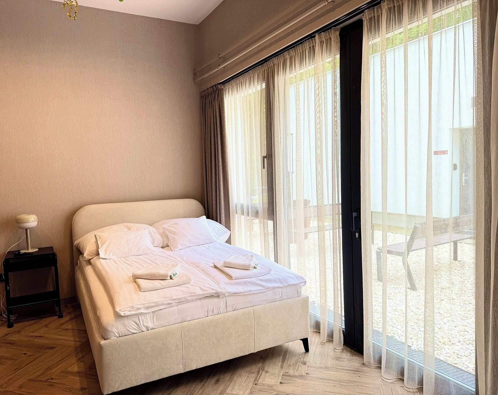 Un dormitorio con cama, mesita de noche y vistas a una zona arenosa a través de amplias ventanas.