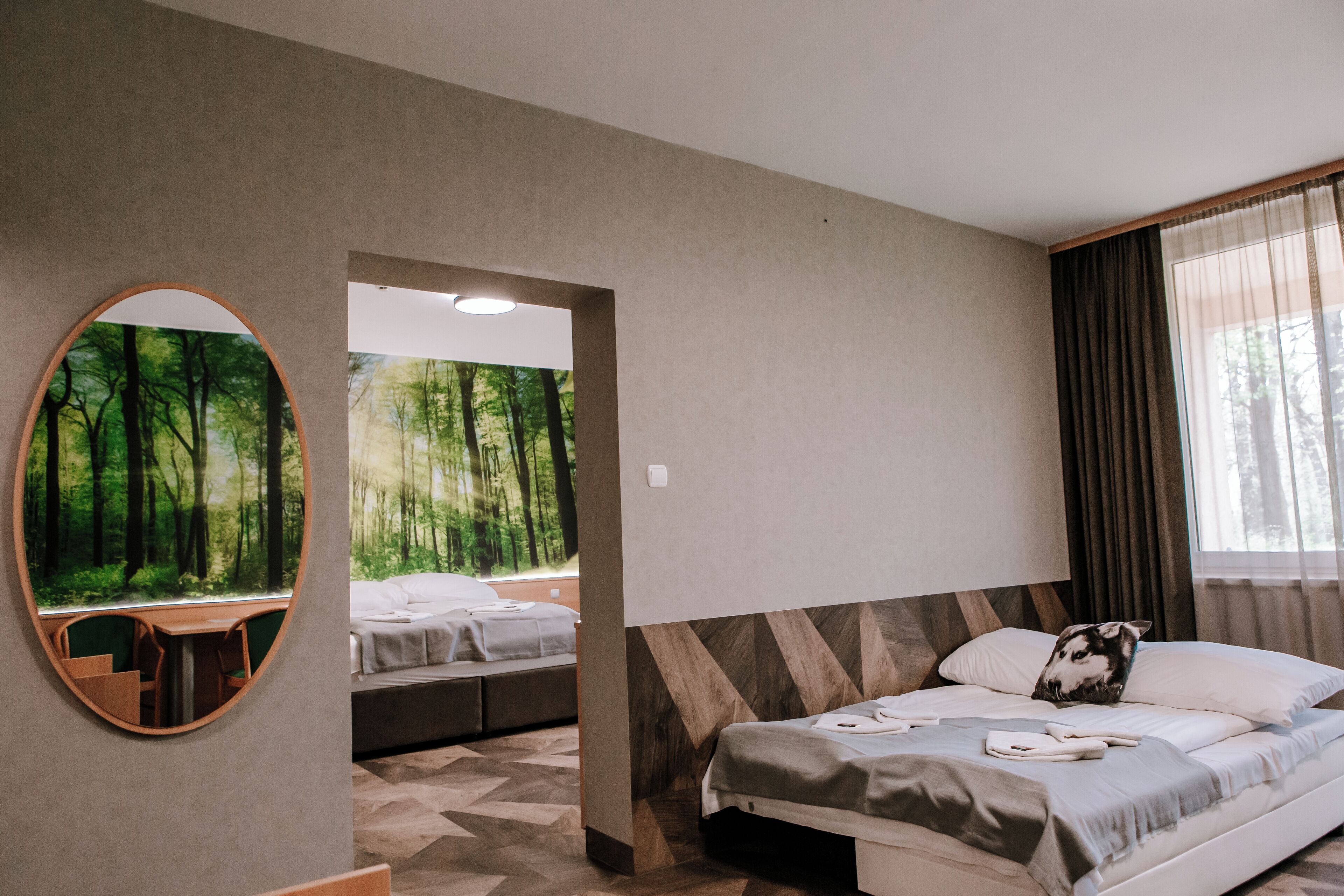 Un dormitorio con cama, espejo, zona de comedor y vista a un bosque por la ventana.