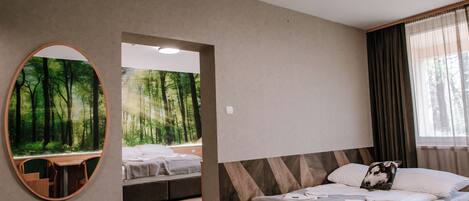 Un dormitorio con cama, espejo, zona de comedor y vista a un bosque por la ventana.