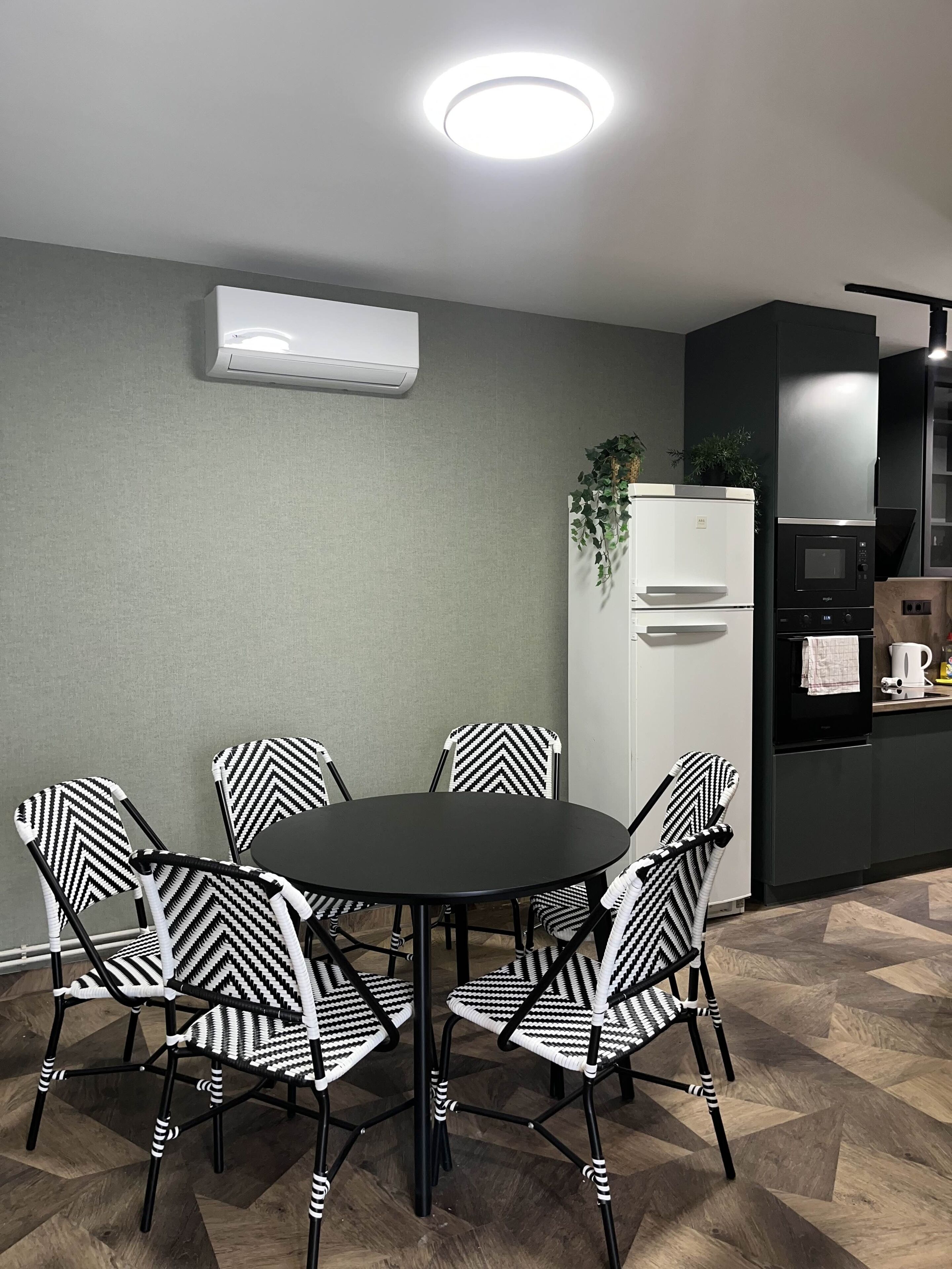 A modern kitchen with a black refrigerator, a microwave, and a coffee maker. There is a dining table with six chairs, each with a black and white patterned seat. An air conditioning unit is mounted on the wall above the table.