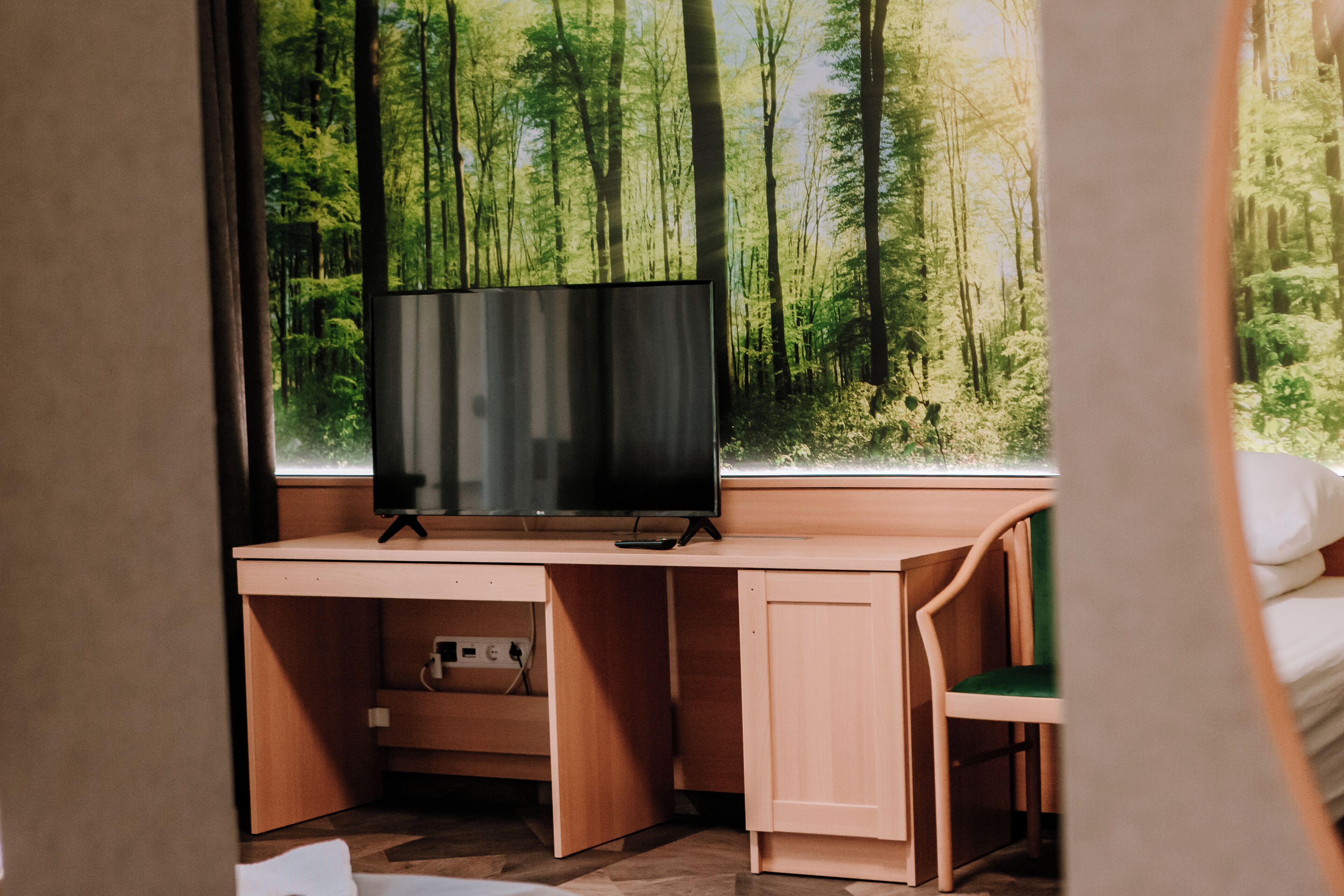 Quarto com escrivaninha de madeira, televisão, cama e janela com vista para uma floresta.
