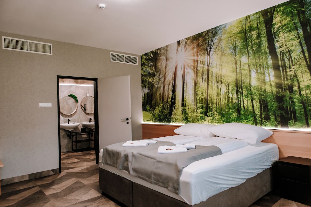 Une chambre avec un grand décor mural représentant une forêt, un lit avec du linge de lit blanc et une salle de bain dotée d’équipements modernes.