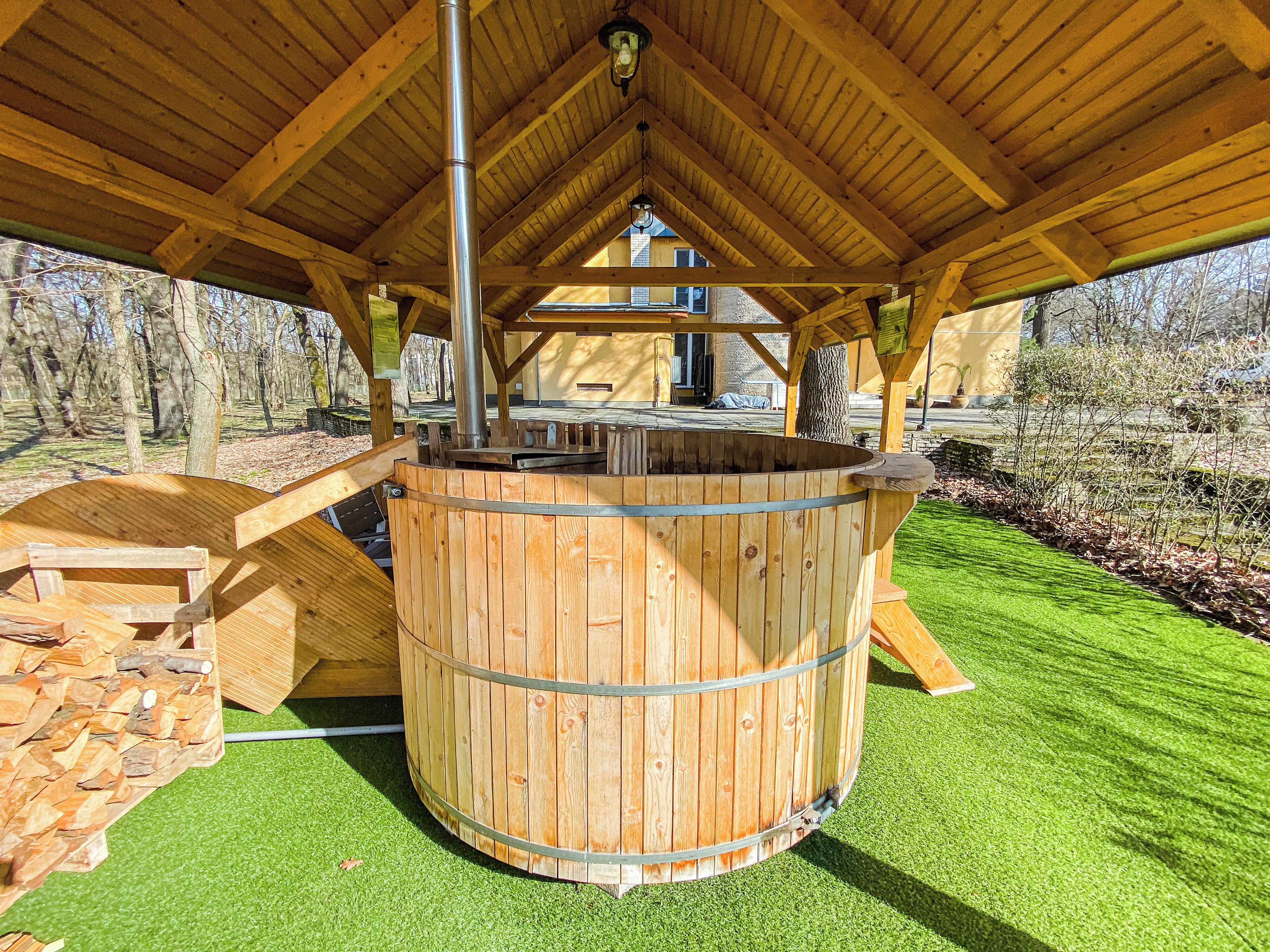 A wooden hot tub under a covered area with a view of a house in the background.