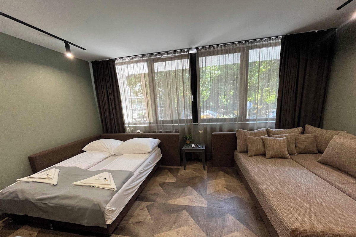 Une chambre d’hôtel avec un lit, un canapé, une petite table et une chaise. Il y a des fenêtres avec des rideaux, et le sol est recouvert d’un motif géométrique.