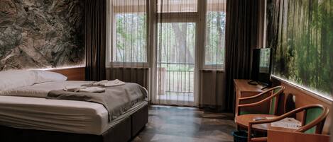 Habitación de hotel con cama, escritorio con sillas, televisor y ventana grande con cortinas.
