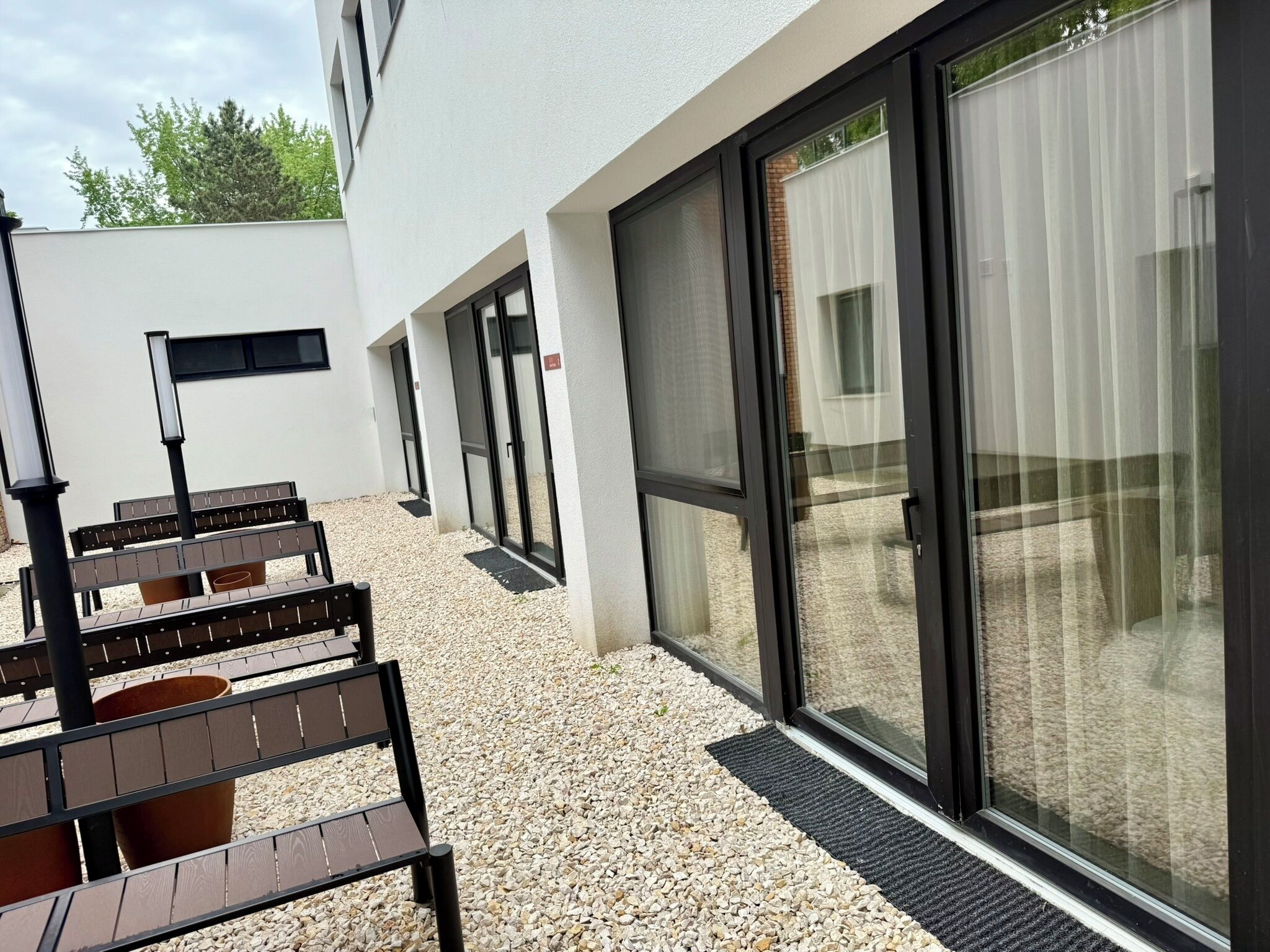 An outdoor seating area with benches and a table, a building with a white exterior, and large glass doors.