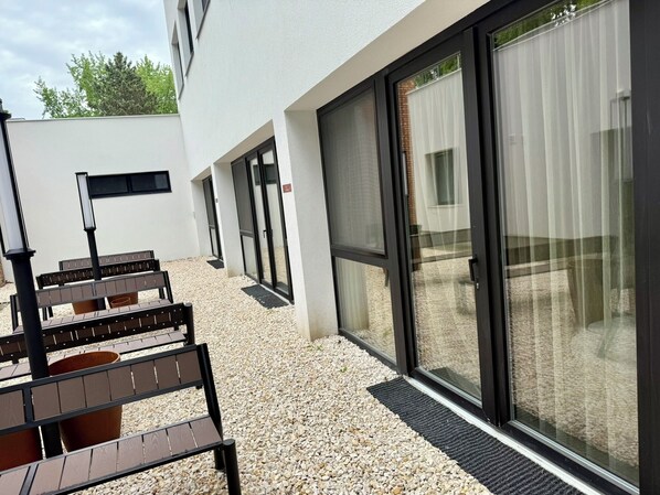 Un espacio al aire libre con bancos y una mesa, un edificio de fachada blanca y grandes puertas de vidrio.