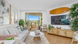 Ein geräumiges Wohnzimmer mit einer großen Couch, einem Couchtisch und einem an der Wand montierten Fernseher. Es gibt einen Holzschrank mit Dekoration und ein Surfbrett, das an der Wand hängt.