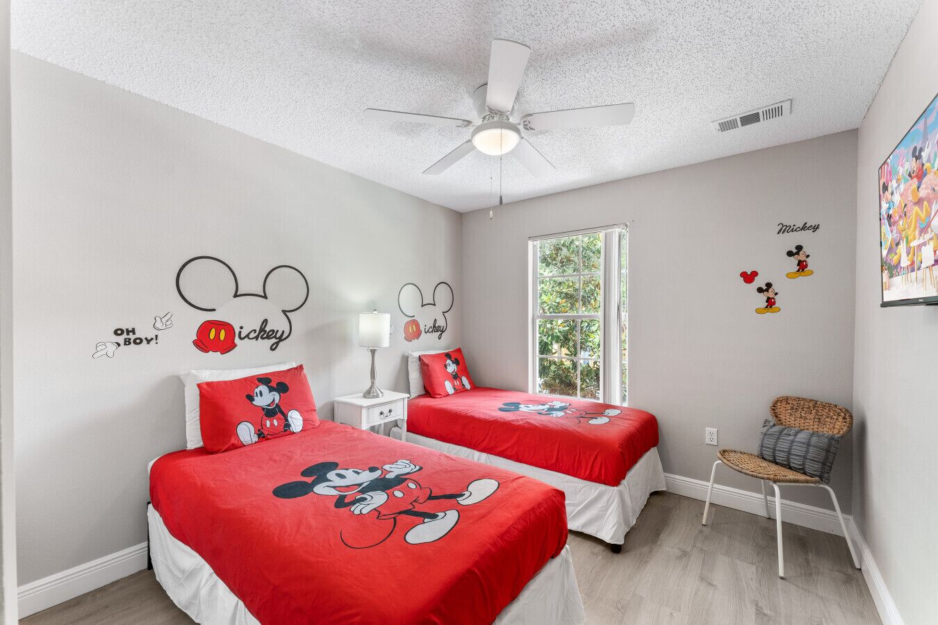 Une chambre avec deux lits, chacun agrémenté de linge de lit aux motifs de Mickey Mouse, un ventilateur de plafond, une fenêtre donnant sur un paysage verdoyant et des autocollants muraux à l’effigie de Mickey Mouse.