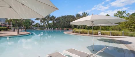 Piscina con sillones de descanso y sombrillas, rodeada de follaje y un cielo despejado.