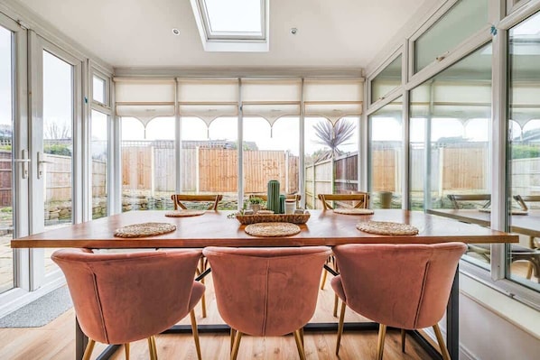 A dining area with a long table, six chairs, and a view of a backyard through large windows.