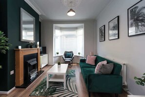 A living room with a fireplace, a green sofa, a white coffee table, and a large window with blinds. - Stylish 3BR House in Hull - Sleeps 5 (Hull)