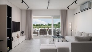 Ein modernes Wohnzimmer mit einer Couch, einem Hocker und einem an der Wand befestigten Flachbildfernseher. Durch die Glastüren ist ein Essbereich mit Stühlen und einem Tisch zu sehen.