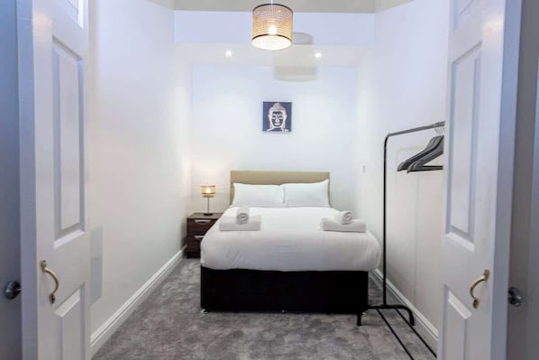 A narrow hotel room with a bed, a nightstand, a wall-mounted lamp, and a Buddha statue. - Cosy & Fully Equipped Cottage in Swinton (Manchester)