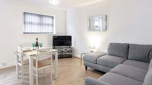 A living room with a grey sofa, a dining table with chairs, a TV, and a window with blinds.