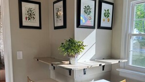 Un coin de pièce avec un comptoir blanc, deux chaises à cadre doré et trois illustrations botaniques encadrées accrochées au mur.