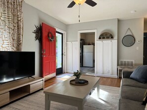 A living room with a flat-screen TV, a coffee table, a red door, and a kitchen in the background. - The Robin's Nest (close to highways, shops and more.) (Greensboro)