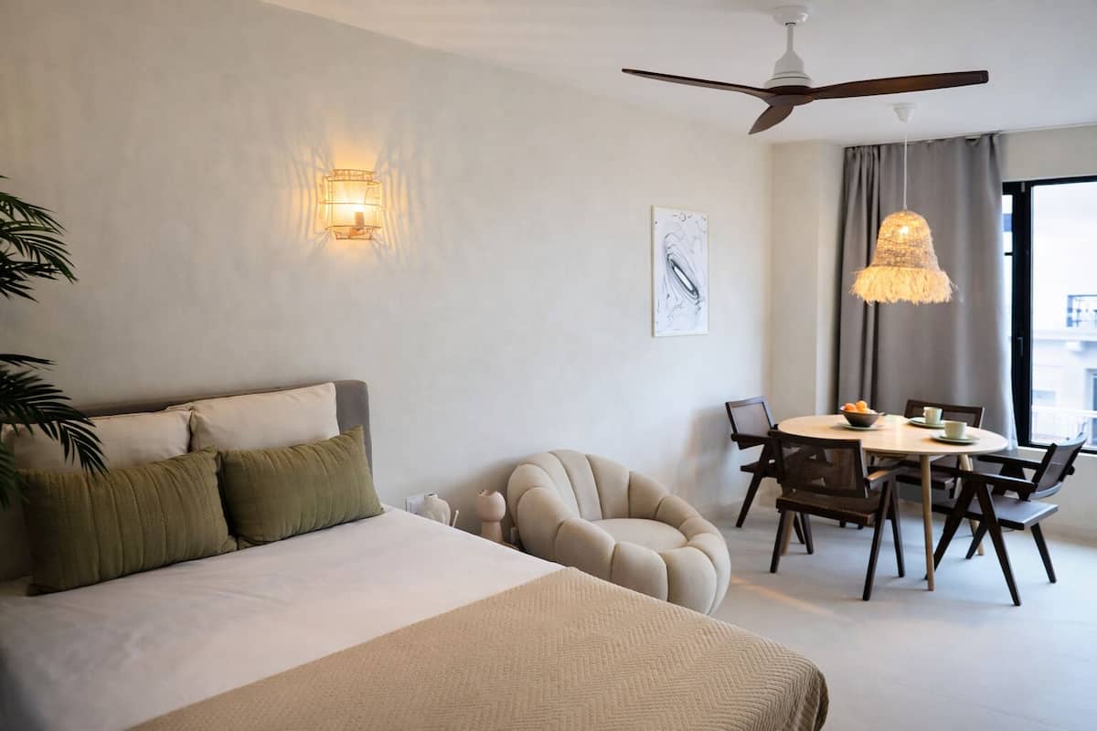 A modern hotel room with a bed, a dining area, a ceiling fan, and a wall-mounted light fixture.