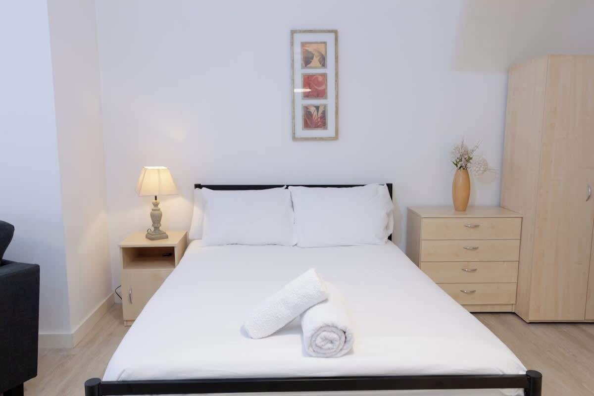 A neatly made bed with white linens, a bedside lamp, a wooden nightstand, a wardrobe, and a framed picture on the wall.