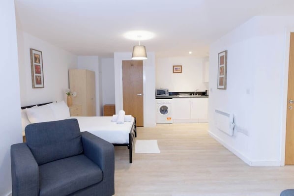 Una habitación de hotel moderna con una cama, un sillón gris, una kitchenette y un armario de madera.