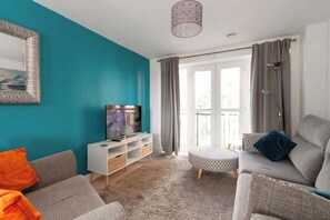 Olohuone, jonka seinät ovat maalattu turkoosilla, siinä on harmaa sohva, valkoinen TV-taso ja suuri ikkuna, jossa on verhot.