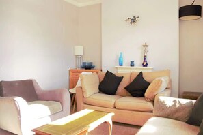 Un salón con un sofá beige, dos sillones, una mesita, una lámpara y una estantería con adornos.