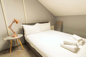 Una cama bien tendida con sábanas blancas, una mesita redonda con una lámpara y un radiador en una habitación con paredes lisas.