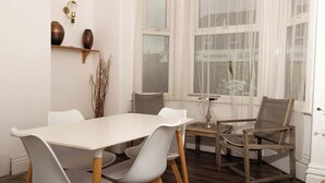 Un comedor con una mesa blanca, sillas grises, una silla de madera, una lámpara y una ventana con cortinas traslúcidas.