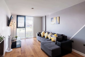 Un salon moderne avec un canapé gris, un radiateur mobile et une grande fenêtre équipée de stores.