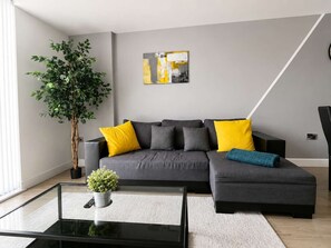 A modern living room with a grey sectional sofa, yellow and grey cushions, a glass coffee table, a potted plant, and a painting on the wall. - Charming 2BR Flat in Sheffield City Centre (Sheffield)