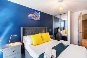 A hotel room with a large bed, a nightstand, a bedside lamp, a painting on the wall, and a door leading to another room. - Lovely 2BR Apt in Sheffield City Centre (Sheffield)