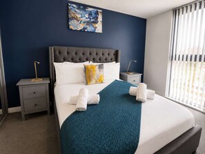 A hotel room with a large bed, a headboard, a nightstand with a lamp, a window with blinds, and a painting on the wall.