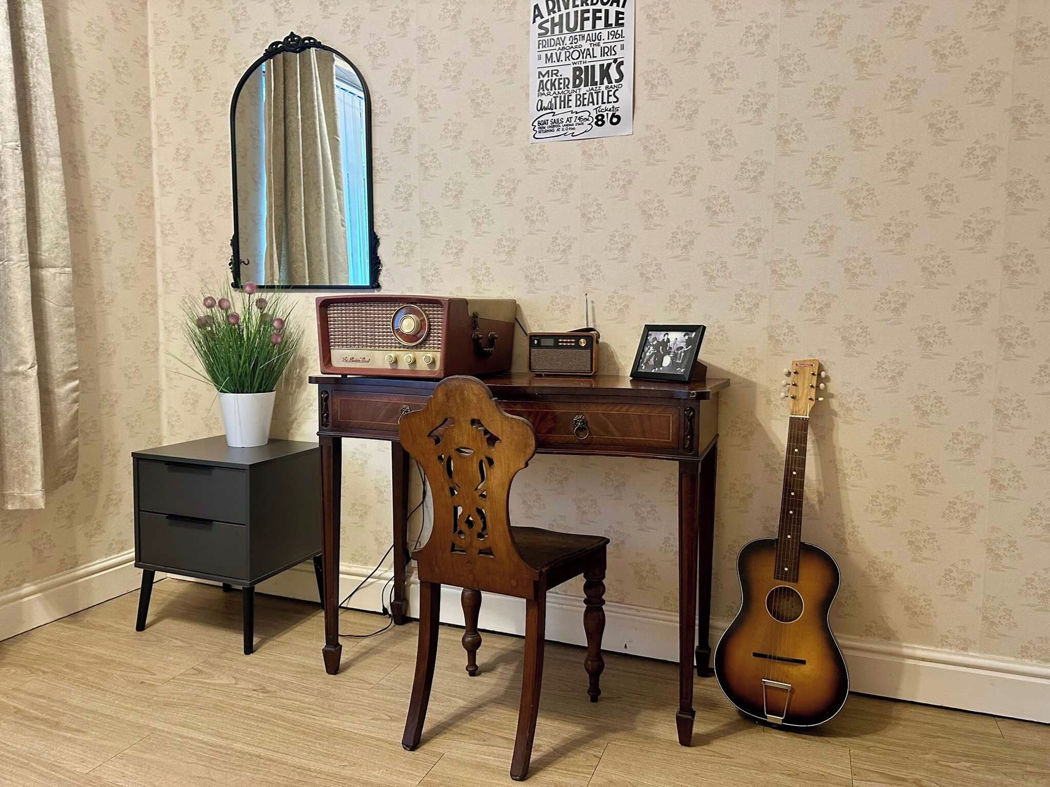 Ein Zimmer mit einem Holztisch, einem Stuhl, einer Gitarre, einem Radio und einem Spiegel.