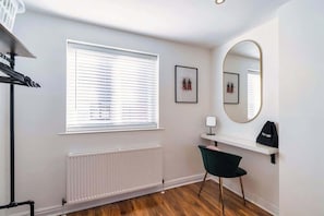 A room with a radiator, a window with blinds, a desk with a chair, a mirror, and a shelf with a bag.