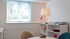 Una habitación moderna con un radiador blanco, una mesa con un frutero, una lámpara con un flamenco y una ventana con persianas.