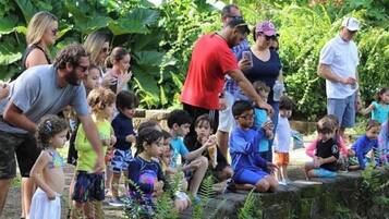 A group of people, including children, gathered outdoors in a lush, green environment with dense foliage and a stone wall.