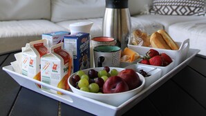 Een ontbijfwagen met melkpakken, sap, fruit en brood op een tafel in een kamer met een bank en decoratieve kussens.