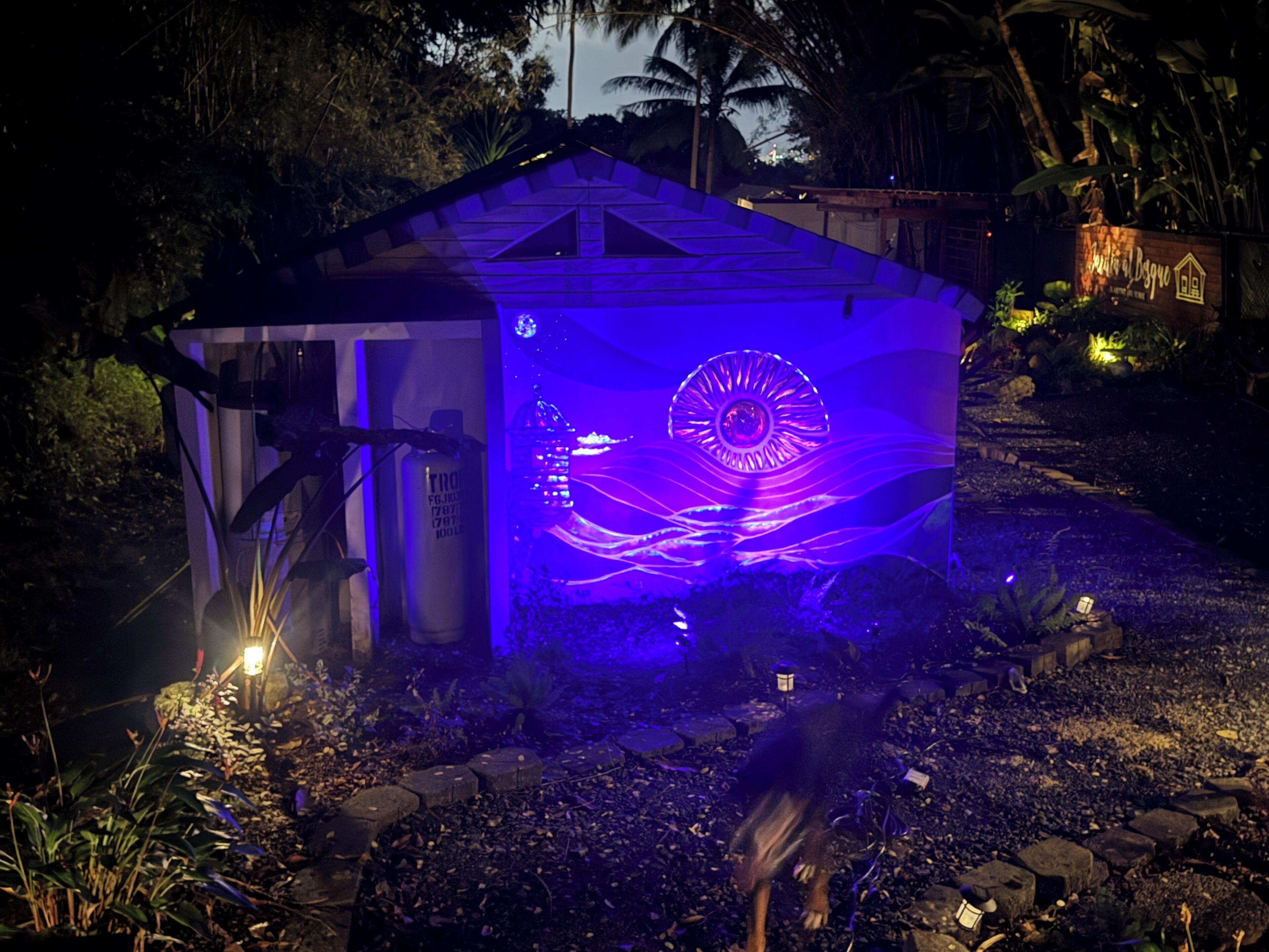 Une petite cabane rustique, dotée d’un éclairage décoratif original sur la façade, entourée d’un jardin où poussent différentes espèces de plantes, et accessible par un sentier.