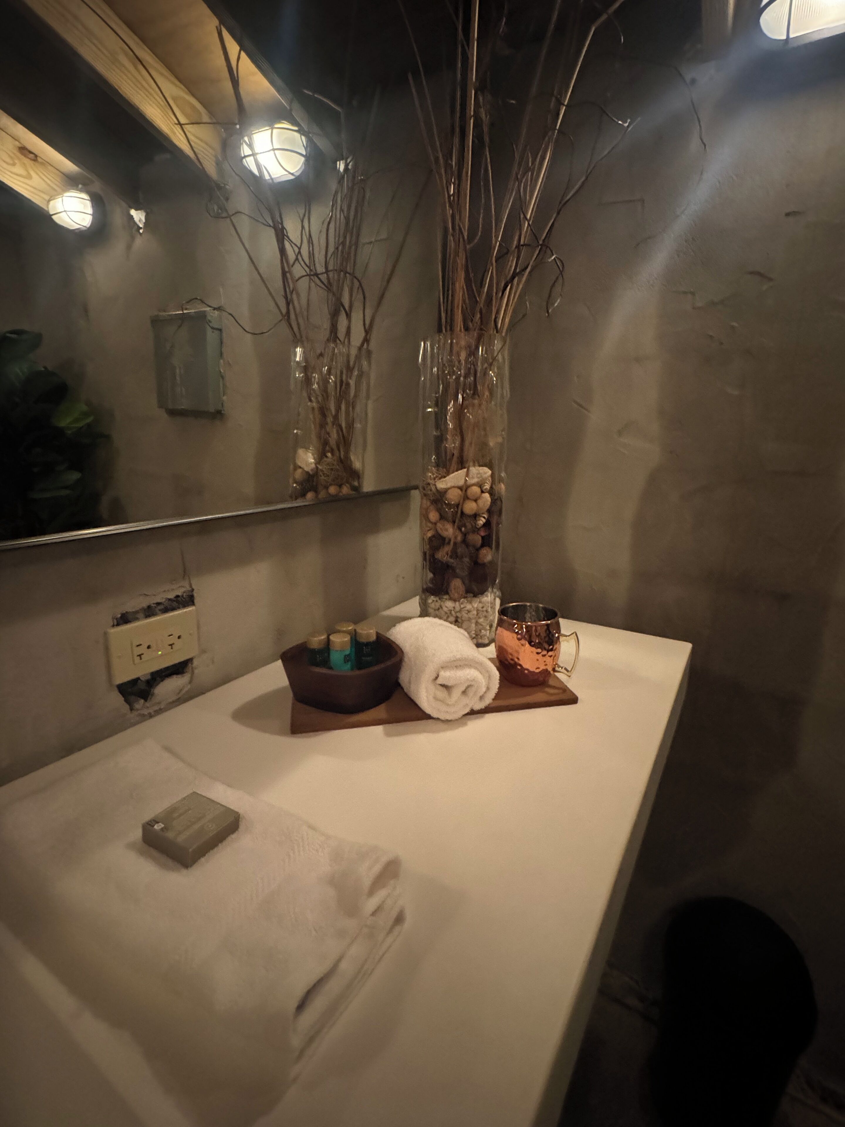 A bathroom with a white countertop, a mirror, a vase with branches and stones, a towel, a copper mug, and a small soap dish.