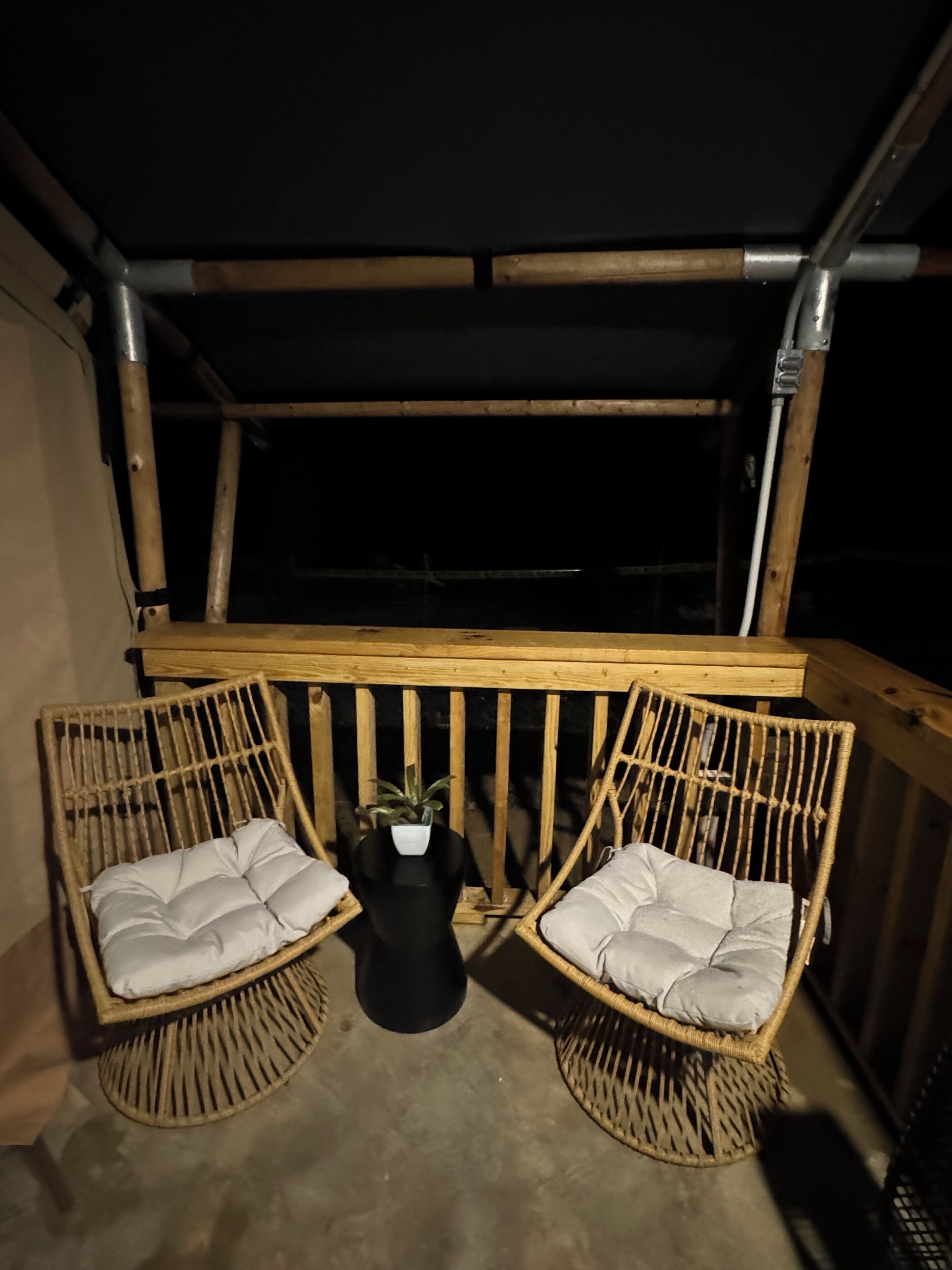 A covered outdoor seating area with two wicker chairs and a small table, under a wooden structure.