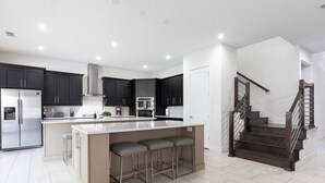 A modern kitchen with dark cabinets, a central island with bar stools, a stainless steel refrigerator, and a staircase with a wooden railing. - Stunning and luxe Bears Den Luxury Villa w/ Outdoor Kitchen near Disney (Kissimmee)