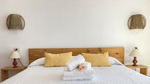 Un lit bien fait, avec du linge de lit blanc, deux oreillers jaunes et une serviette enroulée. Il y a deux tables de chevet avec des lampes, et des décorations murales tissées au-dessus du lit.