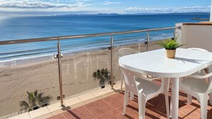 Un balcon avec une table et des chaises blanches, donnant sur une plage d’où l’on a une vue dégagée sur l’océan et les montagnes lointaines.