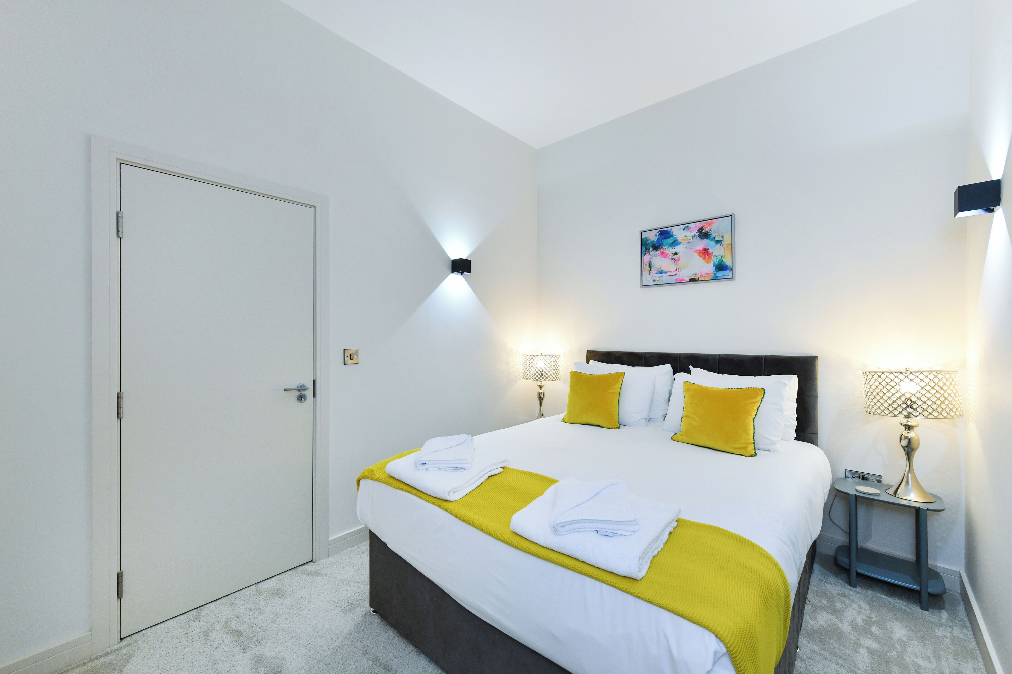 Cama bem arrumada com lençóis brancos e amarelos, mesa de cabeceira com abajur e um quadro na parede.