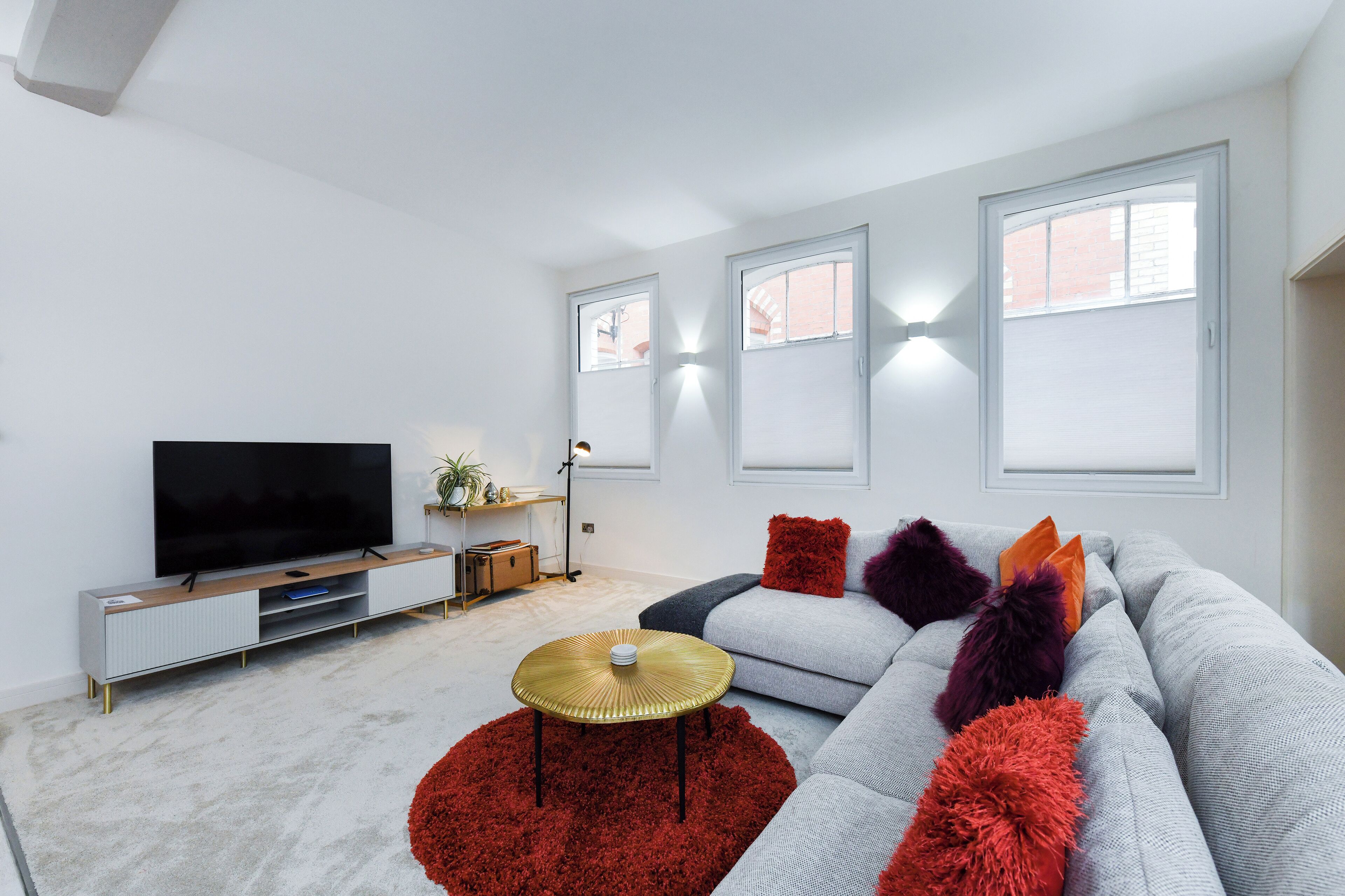 Un salon moderne avec un canapé gris, des coussins de couleur rouge et violette, un téléviseur à écran plat et une petite table ronde surmontée d’un tapis rouge.