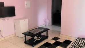 Una habitación con paredes rosas, un refrigerador blanco pequeño, una mesa negra con mantel estampado, una alfombra blanco y negro, y un televisor montado en la pared.