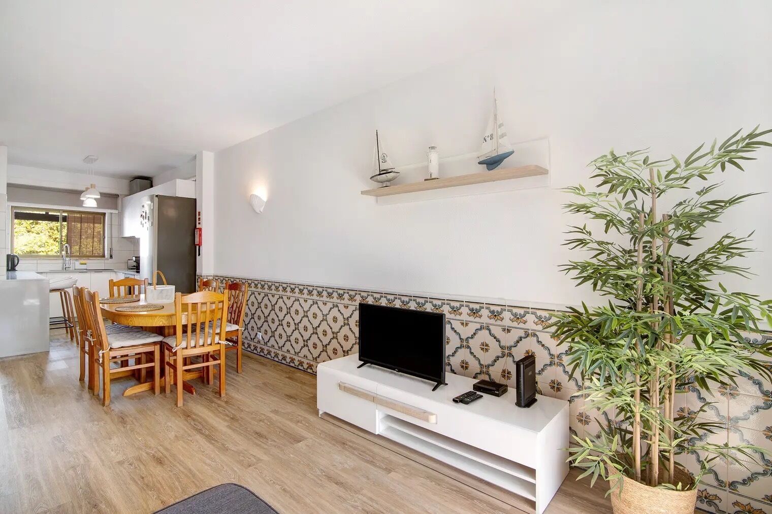 A modern living room with a dining area, a TV on a white console, and a large bamboo plant.