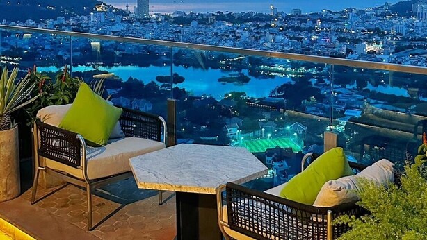 A terrace with wicker chairs and a table, overlooking a cityscape and a body of water at dusk. - 3-bedroom Oasky Vung Tau with Partial Ocean View (Vung Tau)