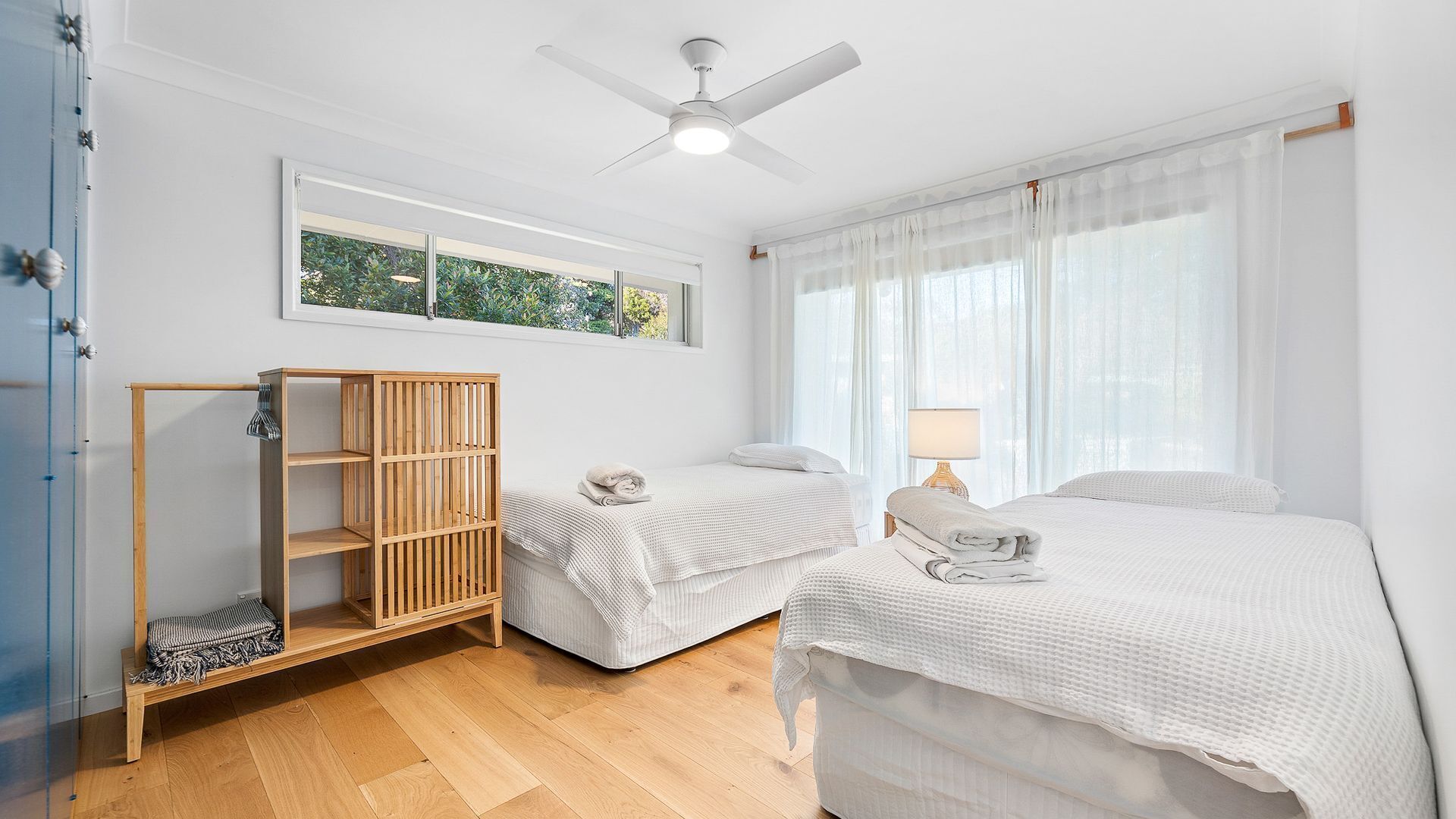Habitación de hotel con dos camas, una estantería de madera, un ventilador de techo y una ventana con cortinas traslúcidas.