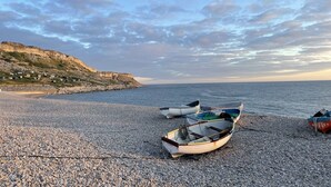 Une plage de galets où plusieurs petits bateaux sont amarrés sur le rivage, une falaise rocheuse en arrière-plan et une mer calme.
